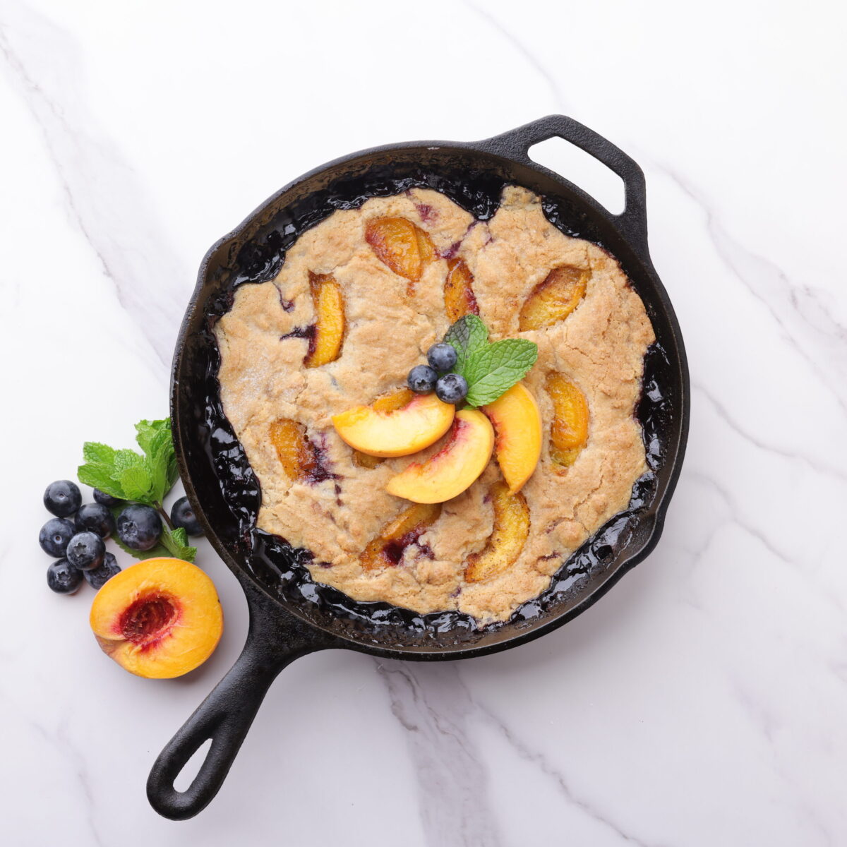 A skillet with the baked blueberry peach cobbler baked to golden perfection, garnished and displayed with blueberries, peaches and mint leaves on top and beside.