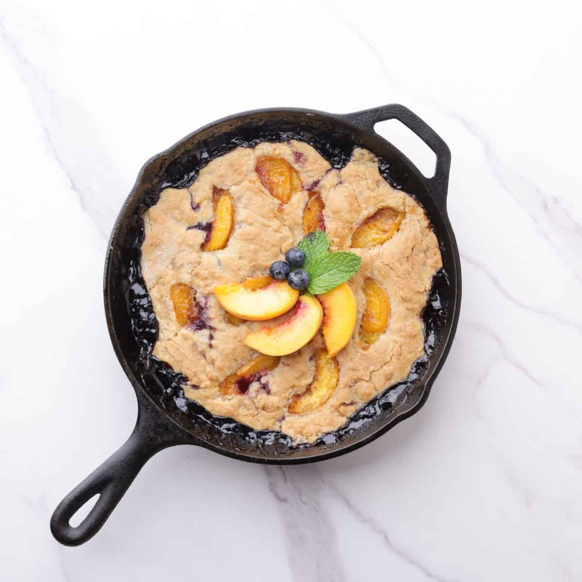 A skillet with the baked blueberry peach cobbler baked to golden perfection, garnished with blueberries, peaches and mint leaves.