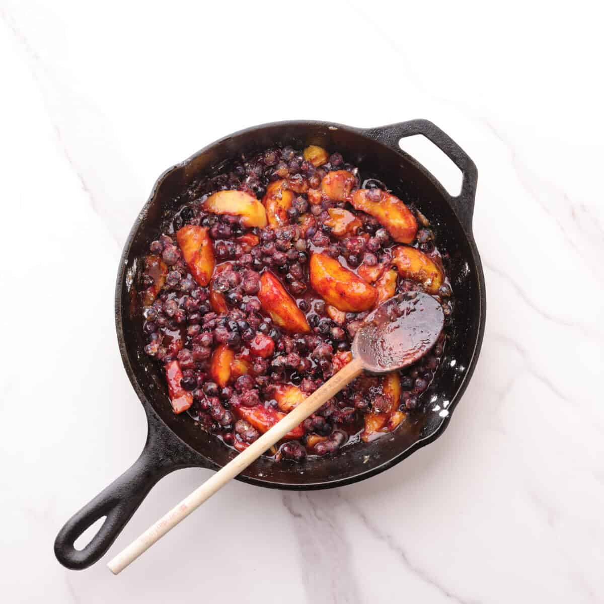A skillet with the blueberries, peaches, spices, sugars and cornstarch combined, displayed with a wooden spoon for stirring.