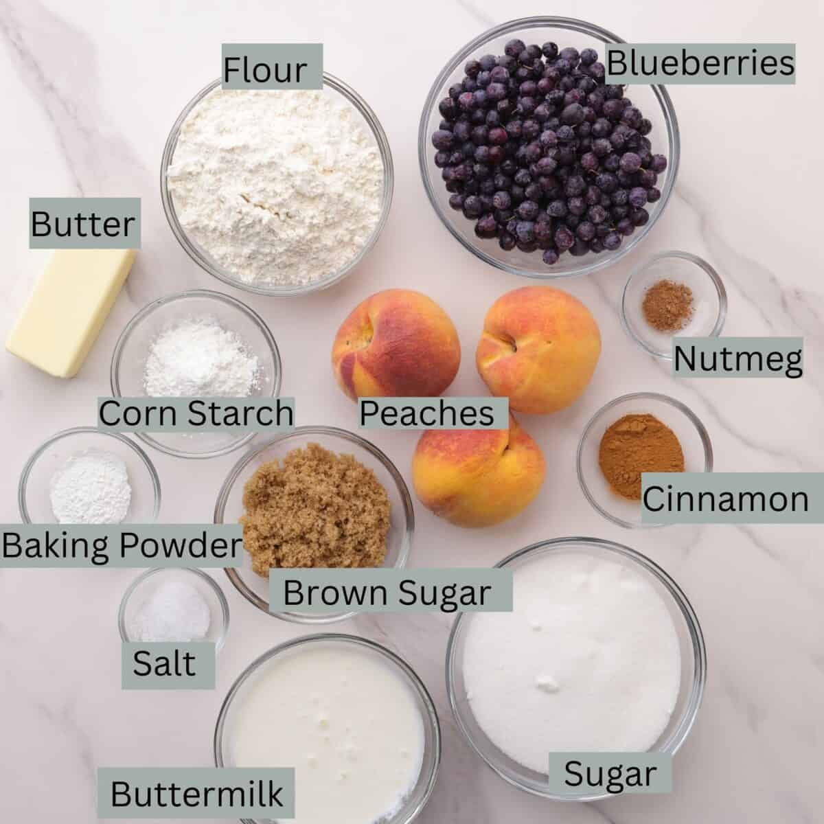 A visual display of the ingredients for the Blueberry Peach Cobbler - wild blueberries, flour, butter, peaches, cornstarch, baking powder, salt, brown sugar, granulated sugar, cinnamon, nutmeg and buttermilk - each in their own bowl.