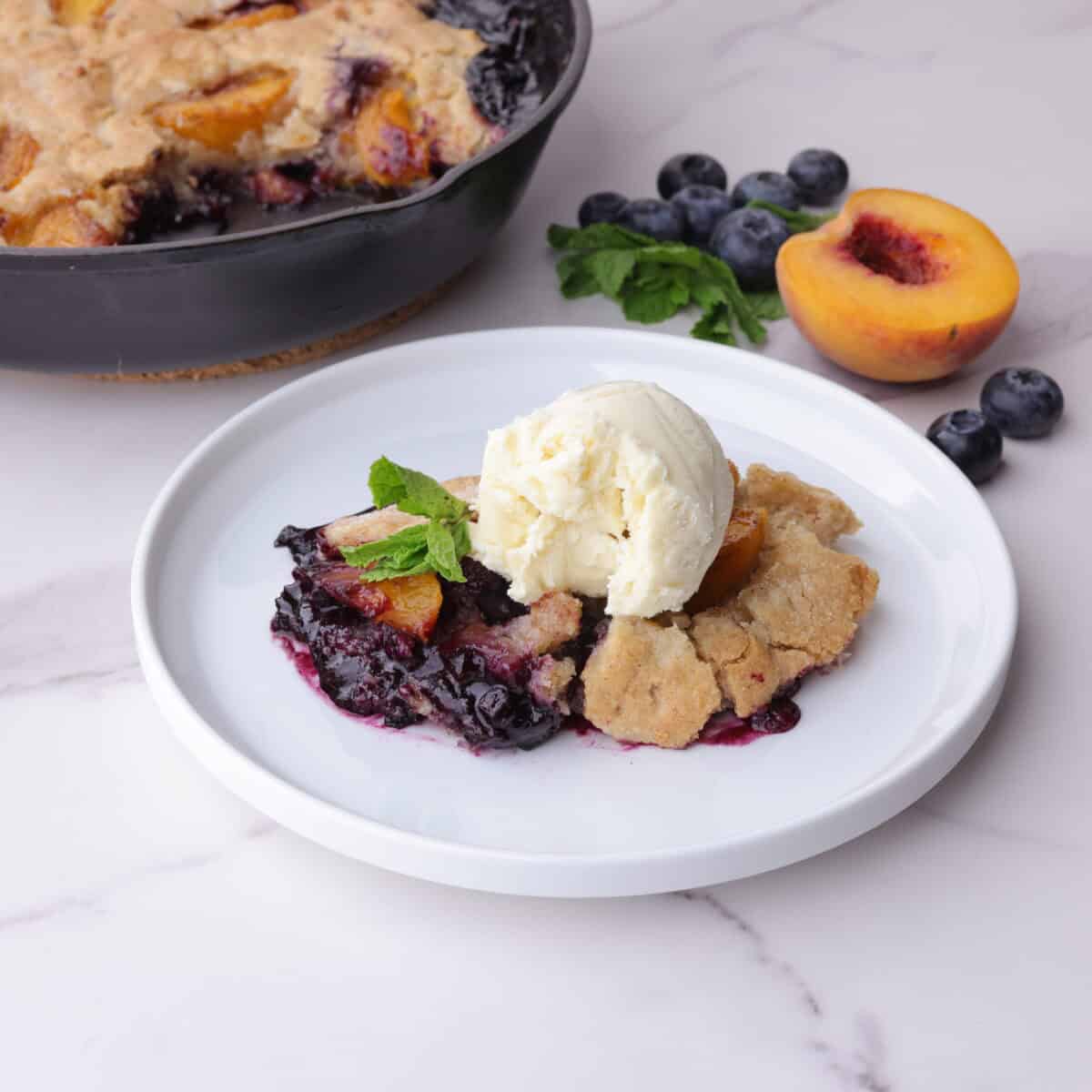 Blueberry Peach Cobbler displayed on a white plate and garnished with mint leaves and ice cream with mint, blueberries and a half peach beside it and the remaining cobbler in the skillet in the background.