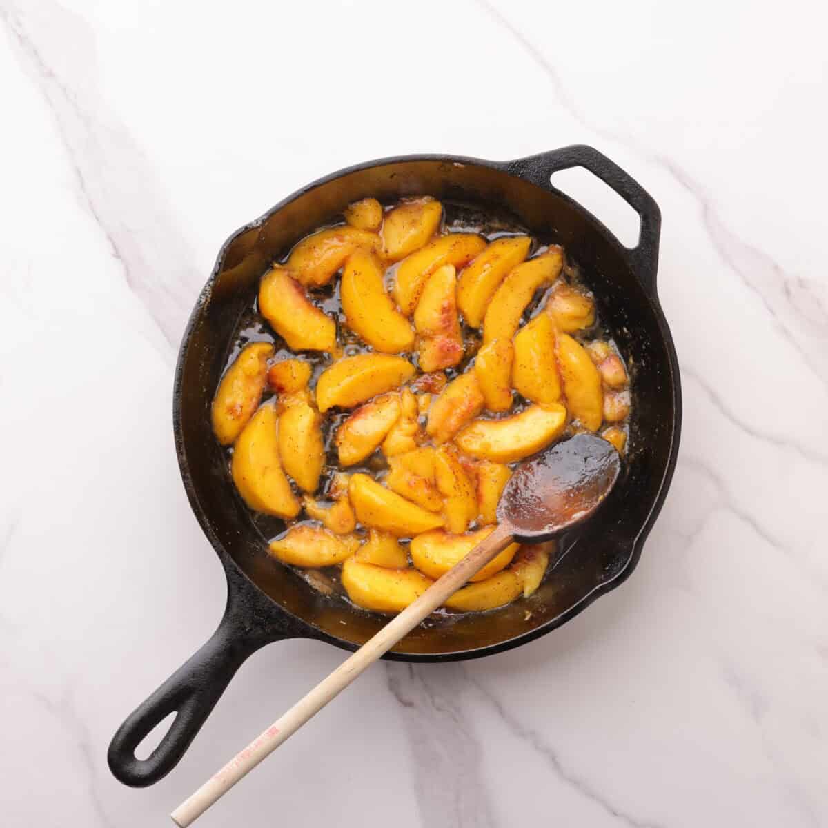 A skillet with golden peaches sautéd in the browned butter, displayed with a wooden spoon for stirring.
