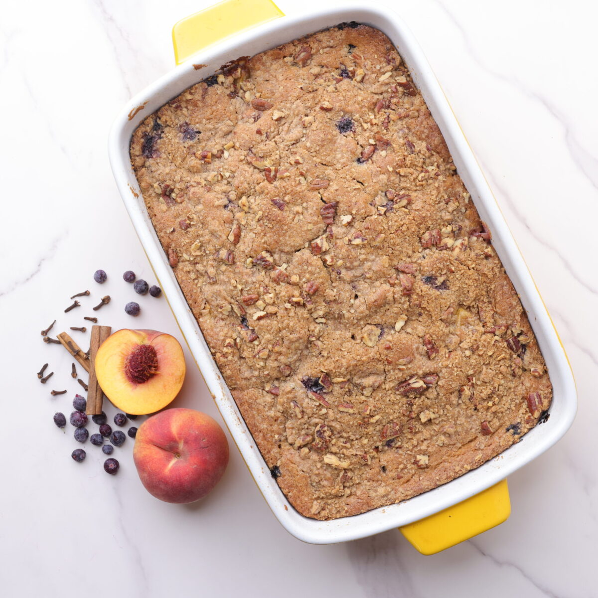 A beautifully golden brown baked Peach and Blueberry Coffee Cake in the baking dish displayed with peaches, blueberries, cinnamon sticks and cloves arranged beside. 