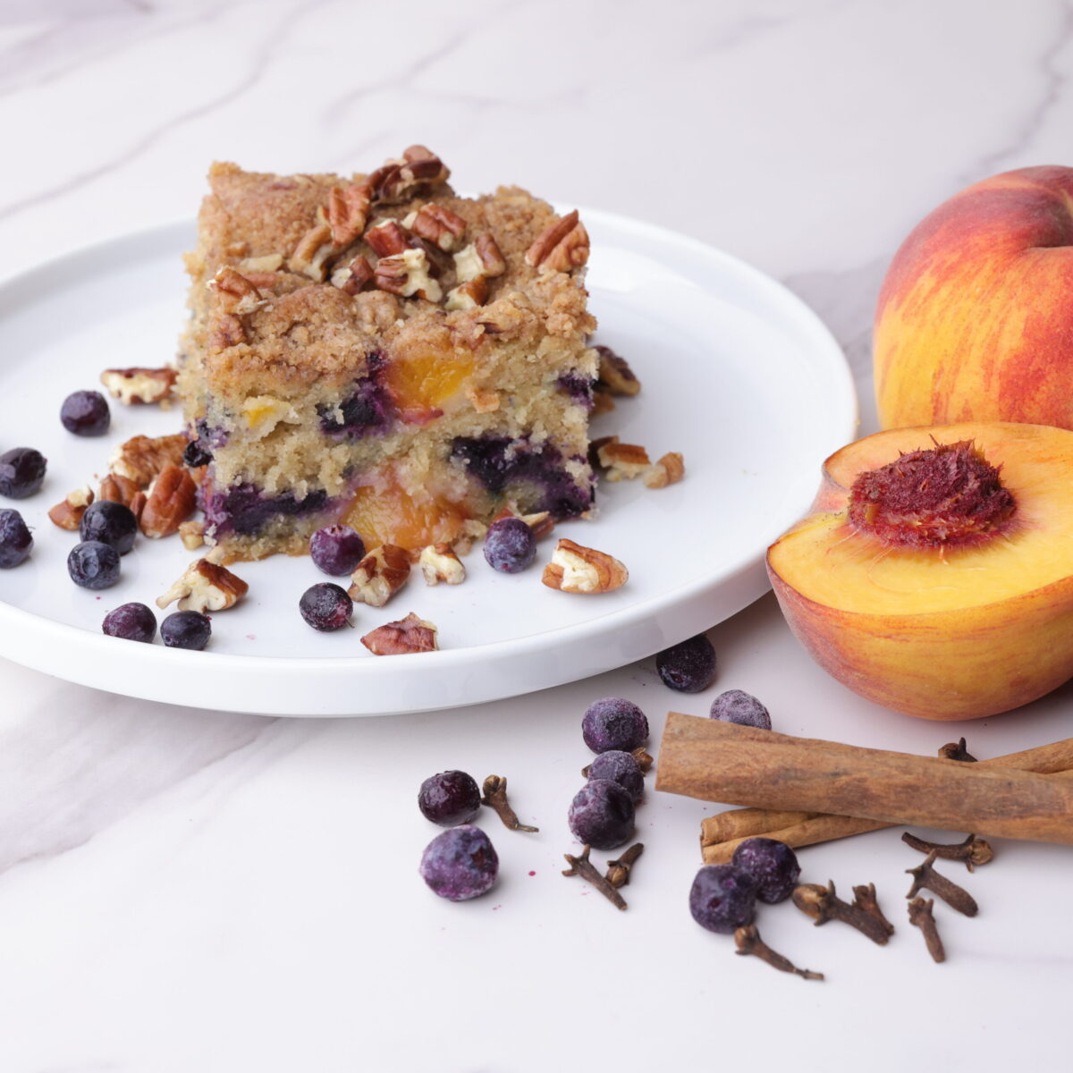 A slice of Peach and Blueberry Coffee Cake displayed on a white plate with peaches, blueberries and cinnamon sticks beside.