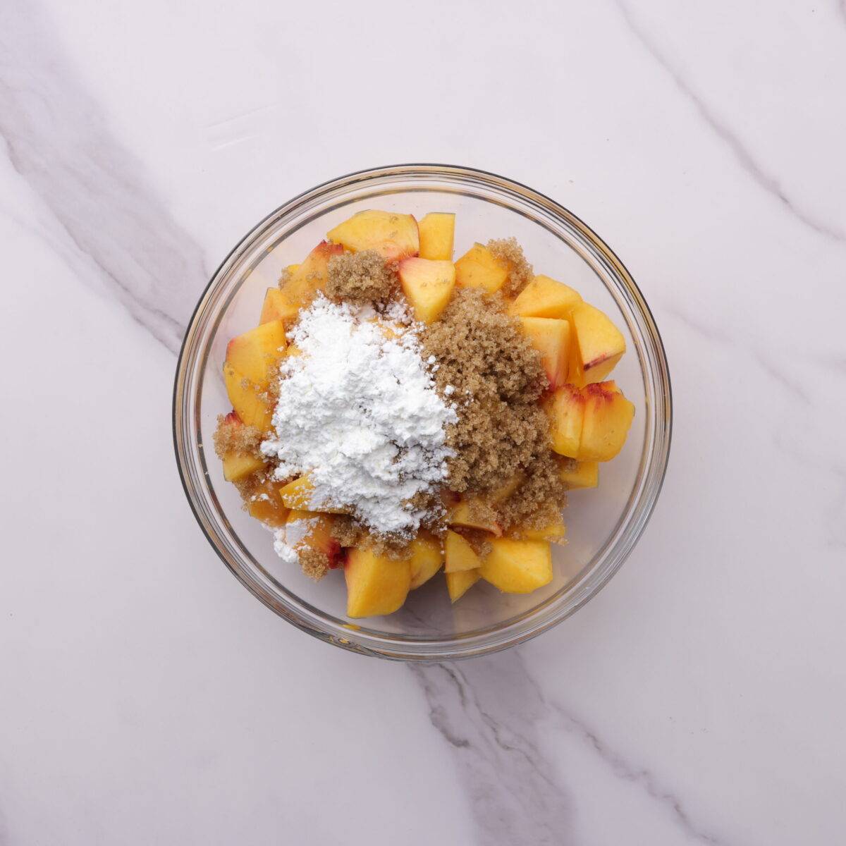 A glass bowl with the diced peaches topped with brown sugar and cornstarch, ready to be combined. 