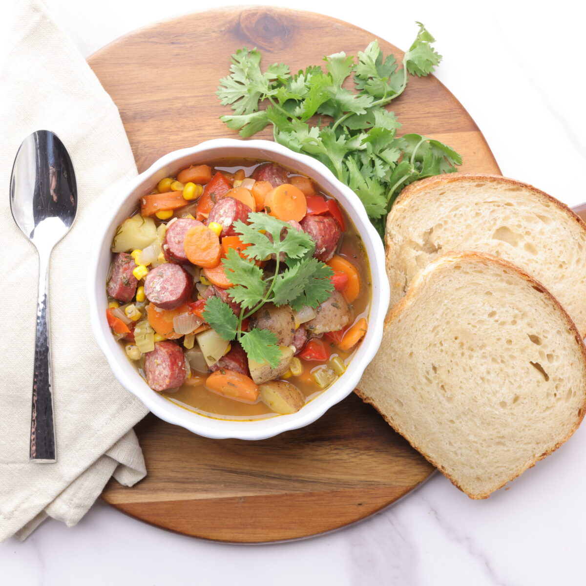 A bowl of Sausage and Pepper Soup displayed on a wooden board with bread, parsley and a spoon on a cloth napkin.