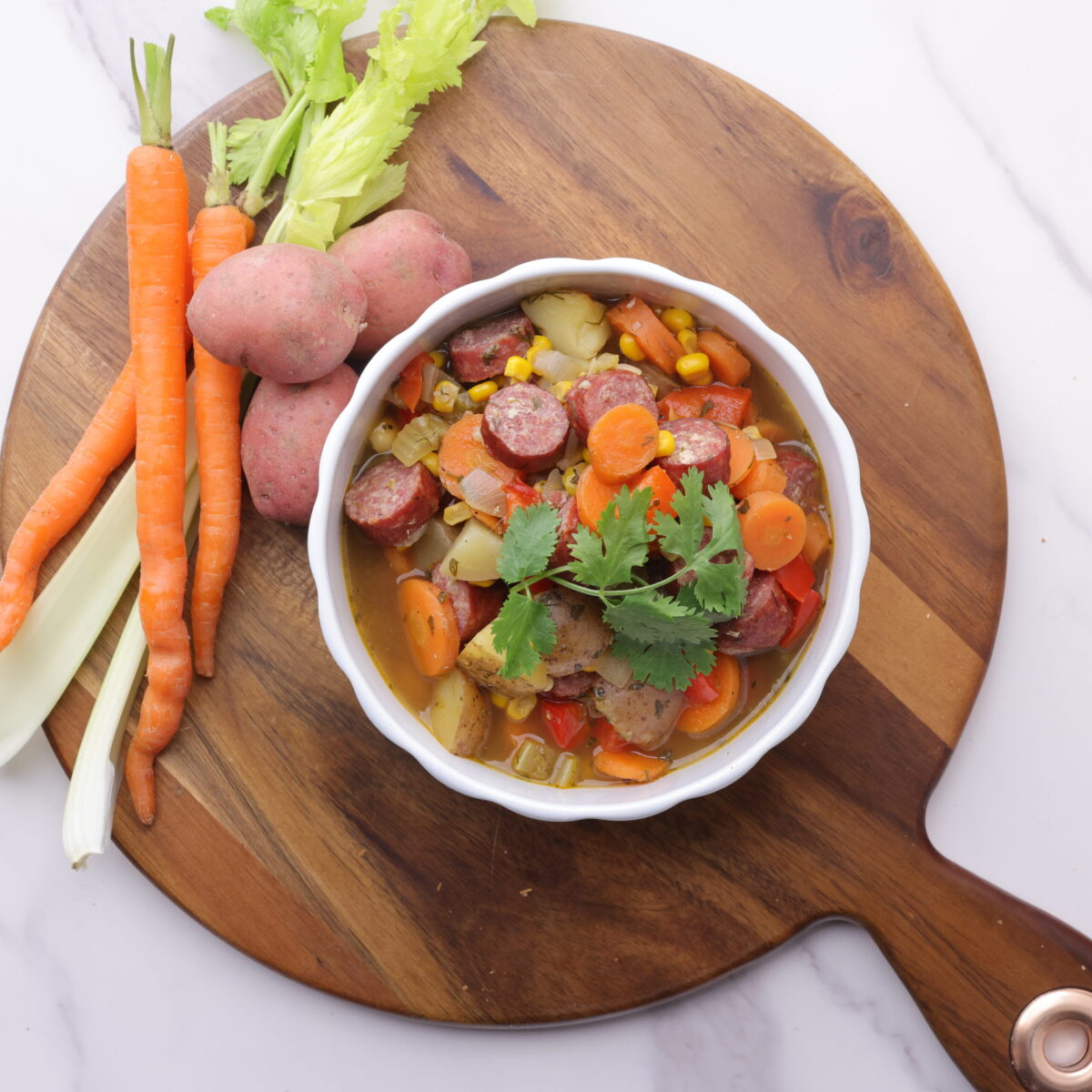 A bowl of Sausage and Pepper Soup displayed on a wooden board with carrots, celery and potatoes beside it.