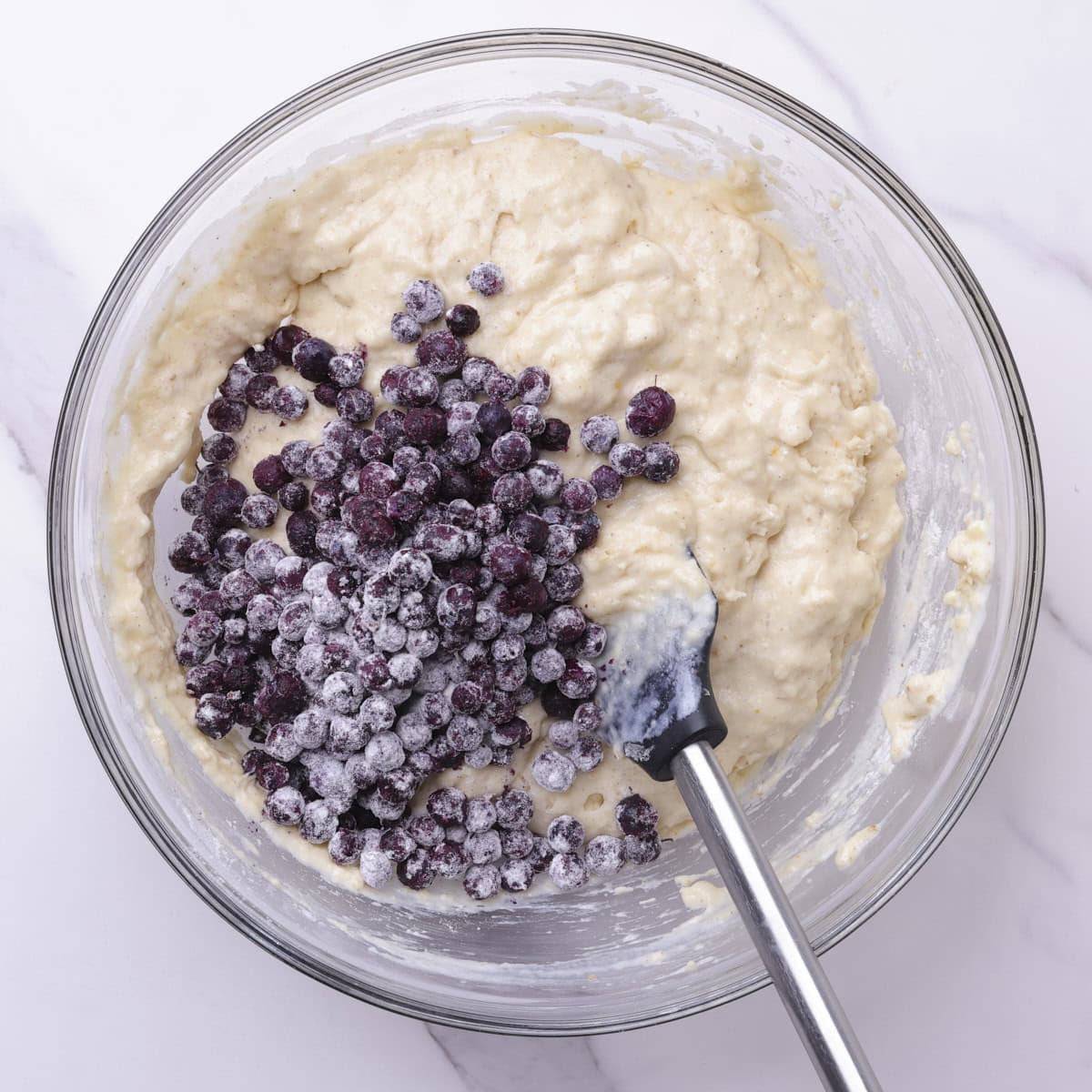 The muffin batter with the floured blueberries resting on top with a spatula ready to fold them in.