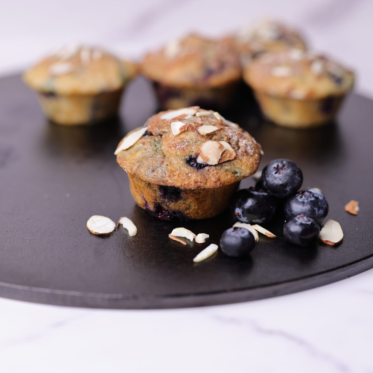The blueberry muffins displayed on a cast iron sheet, one in the foreground displayed with blueberries and slivered almonds.