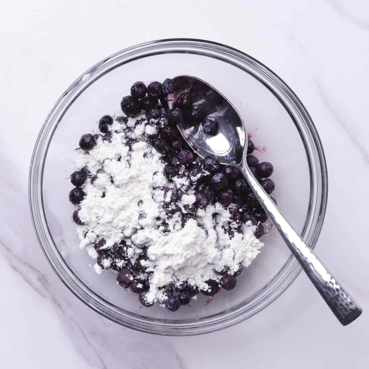 A small glass bowl with the blueberries sprinkled with flour and a spoon for mixing.