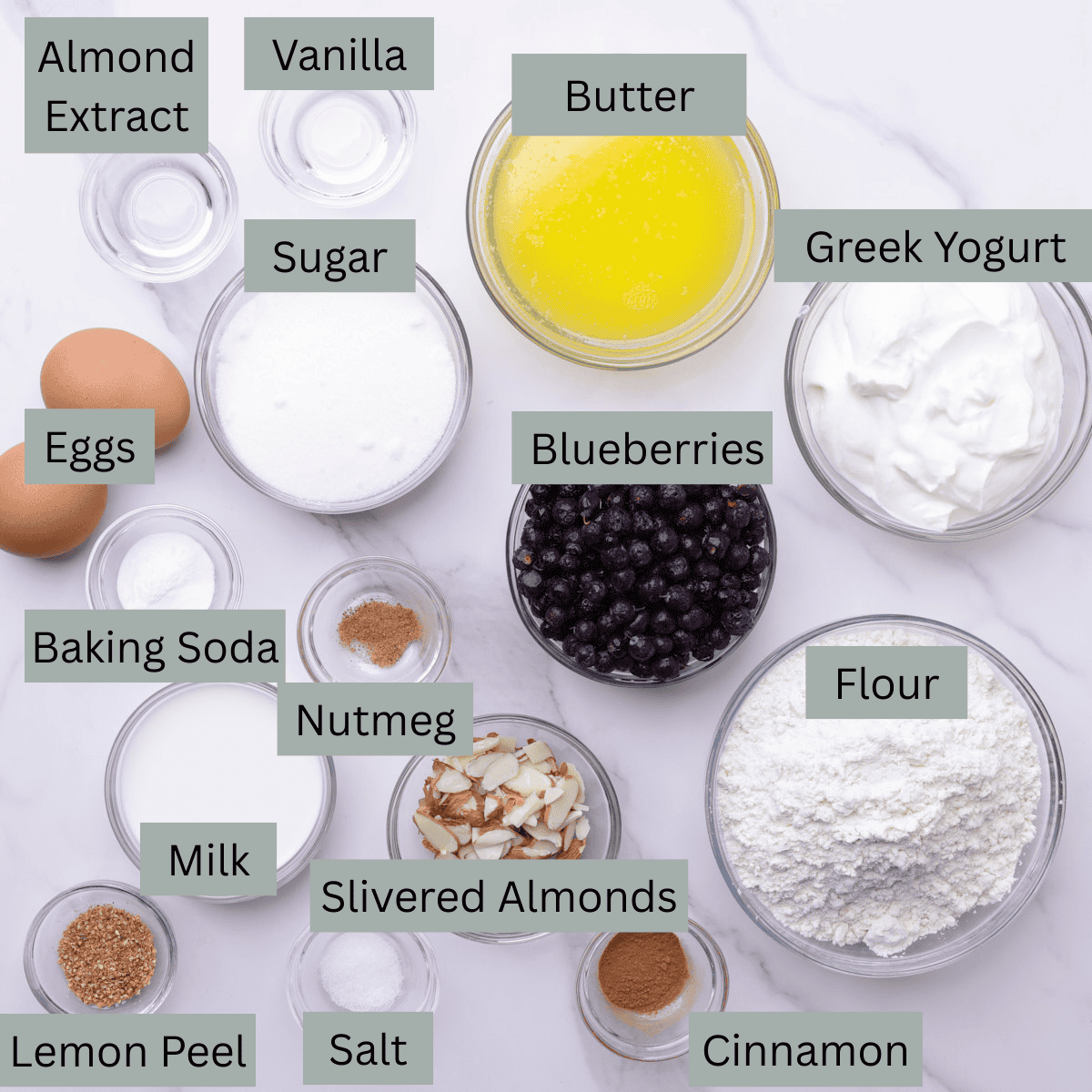 A visual display of the ingredients needed for the Blueberry Muffins with Greek Yogurt - melted butter, Greek yogurt, blueberries, flour, cinnamon, salt, lemon peel, nutmeg, baking soda, sugar, milk, slivered almonds, eggs, vanilla and almond extract - each in their own bowl.