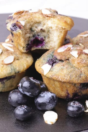 Blueberry Muffins using Greek Yogurt stacked with one bitten muffin on top, garnished with blueberries and slivered almonds.
