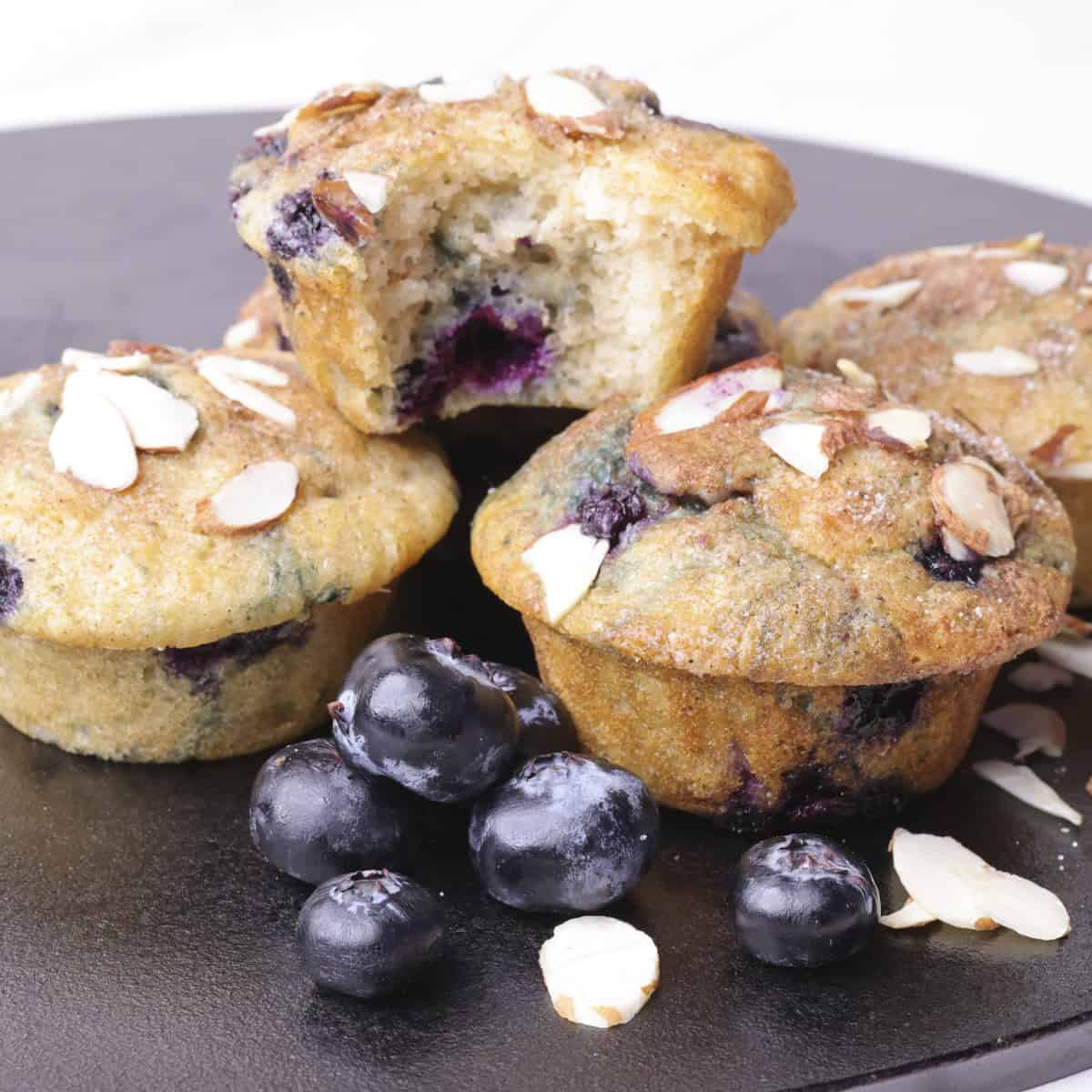 Blueberry Muffins using Greek Yogurt stacked with one bitten muffin on top, garnished with blueberries and slivered almonds.