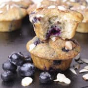 Two Blueberry Muffins using Greek Yogurt stacked with one bitten muffin on top, garnished with blueberries and slivered almonds.