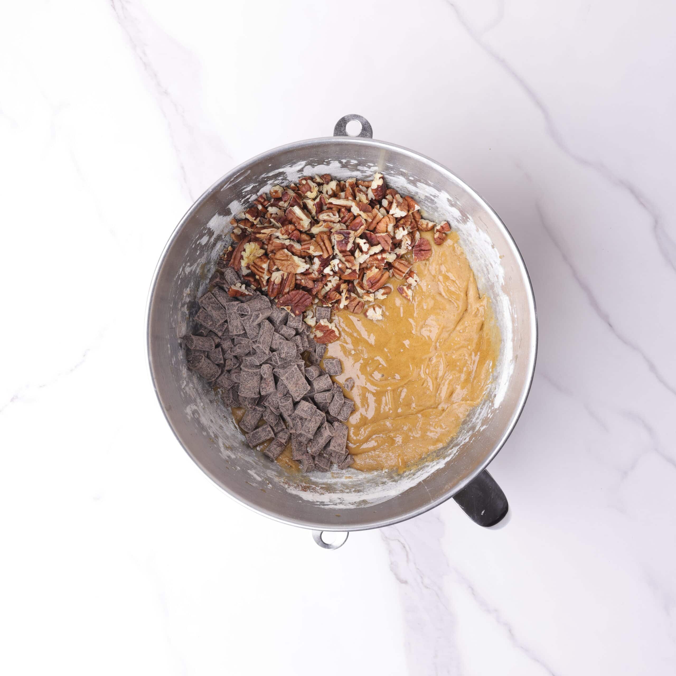 A mixing bowl with the batter in the bottom and the chocolate chunks and pecans sitting on top.