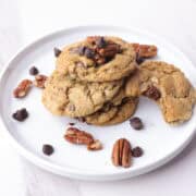 A white plate with a stack of chocolate chunk pecan cookies on it with one bitten cookie on the side, all sprinkled with pecans and chocolate chips.