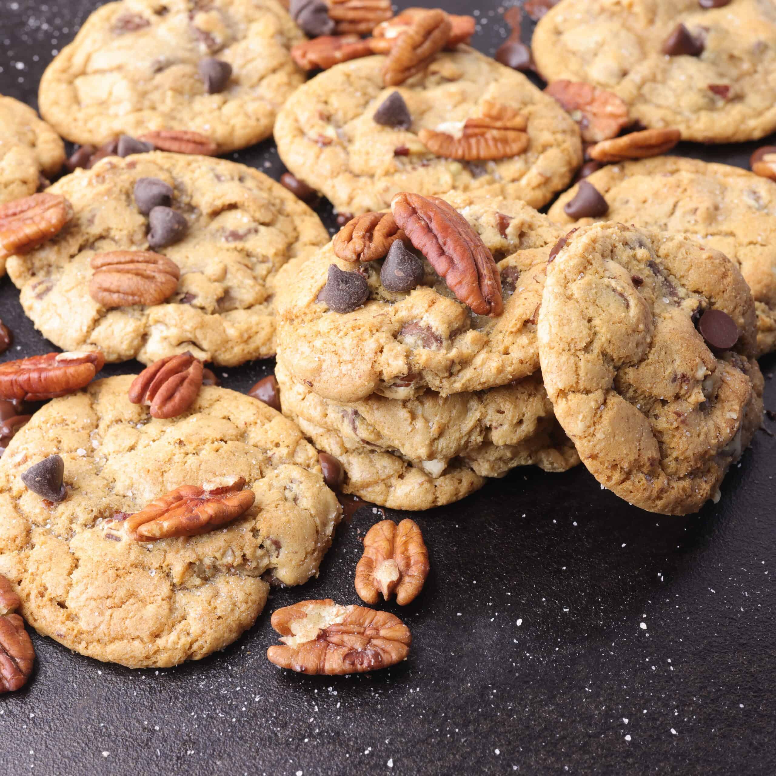 A cast iron baking pan with baked chocolate chunk pecan cookies on it, sprinkled with coarse salt, pecans and chocolate chips.