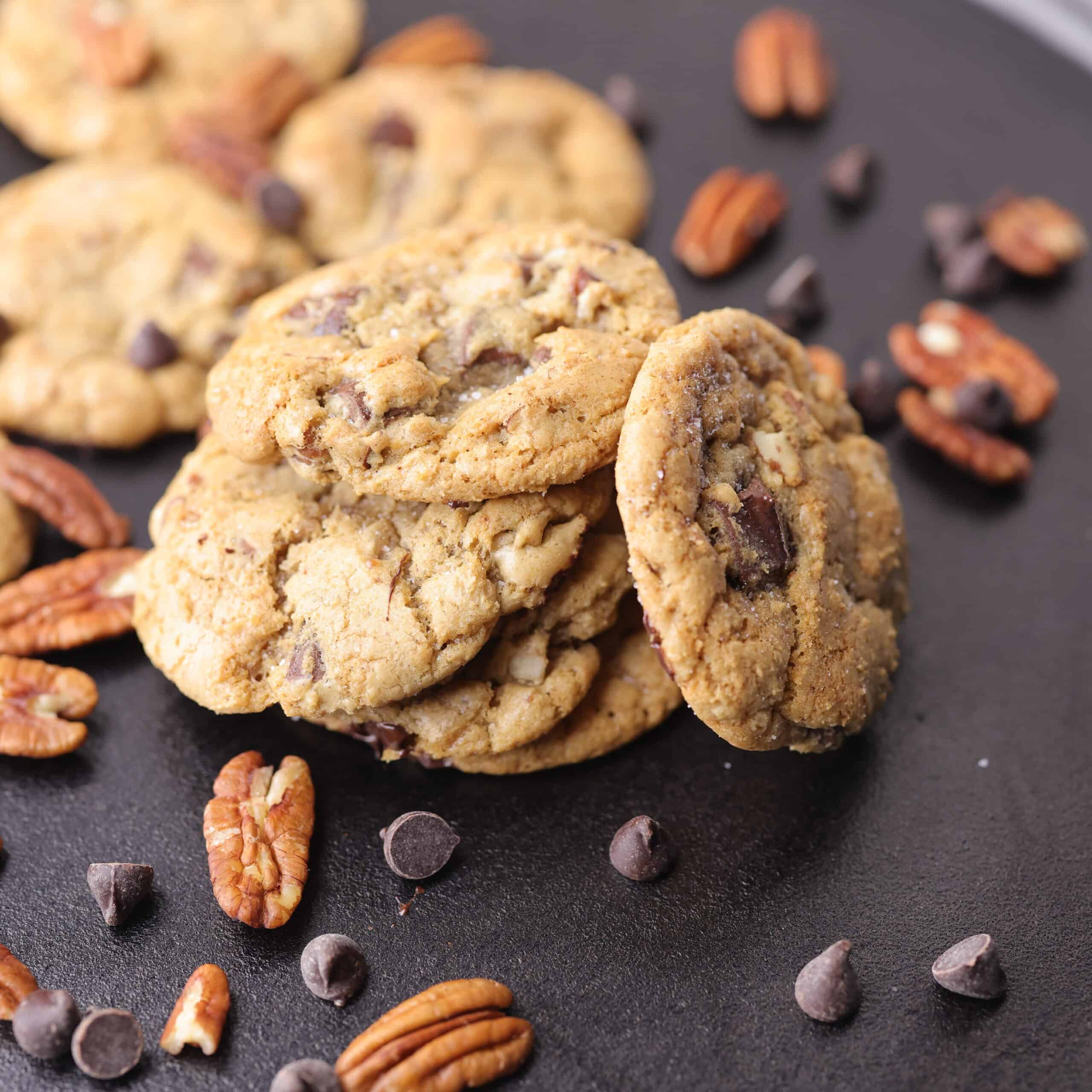 A stack of Chocolate Chunk Pecan cookies on a cast iron plate, sprinkled with sea salt, pecans and dark chocolate chips.