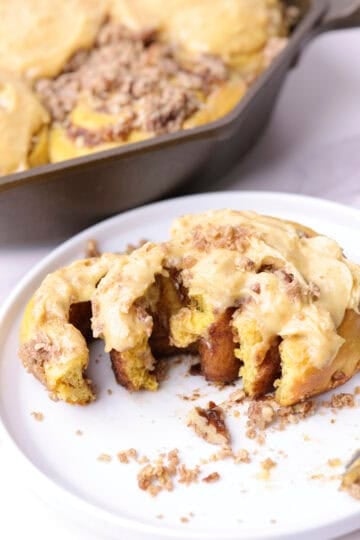 A pumpkin cinnamon roll broken open, displayed on a white plate with a fork and a skillet of rolls in the background.