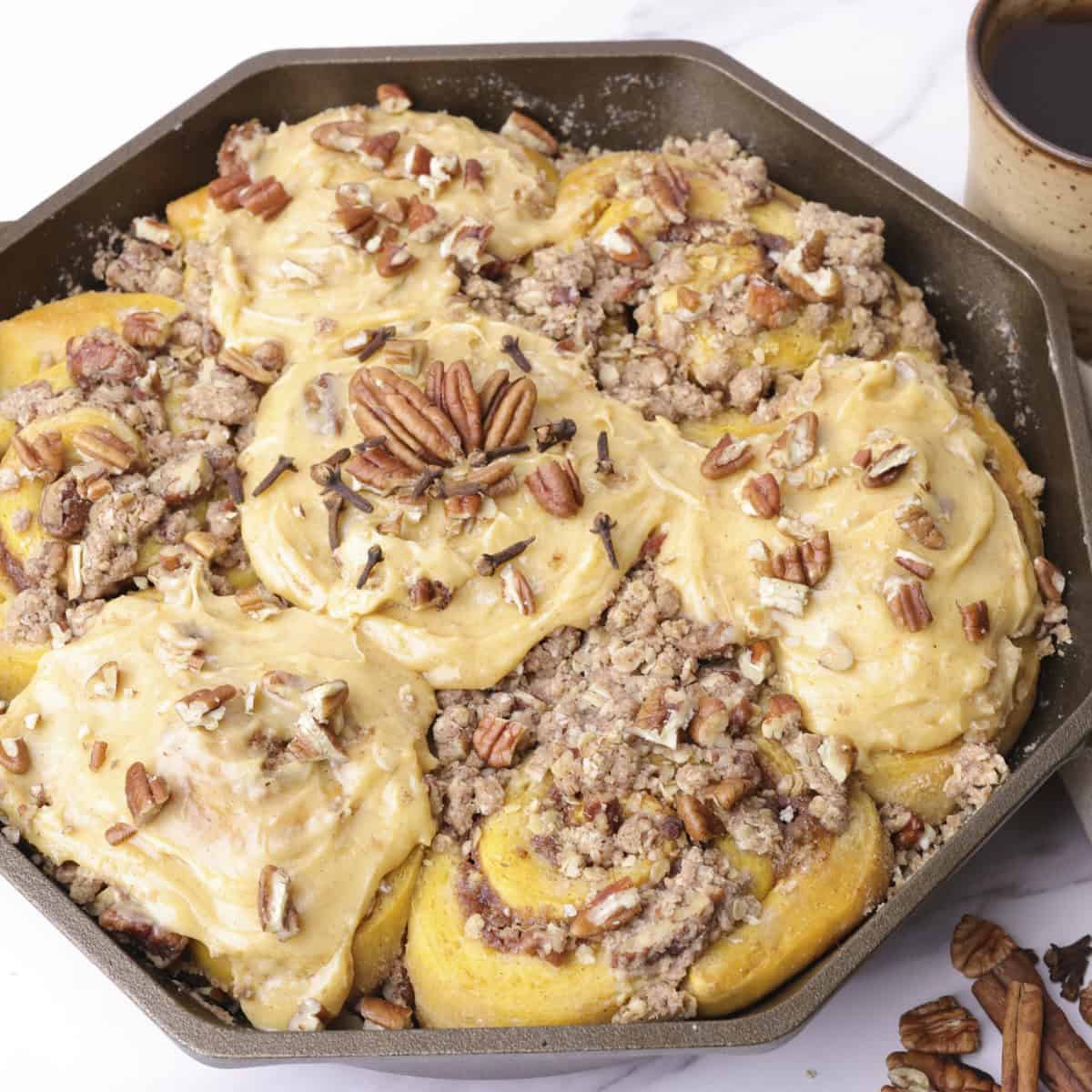 A skillet with the baked pumpkin cinnamon rolls in it with every other topped with pumpkin cream cheese frosting and garnished with pecans and cloves.