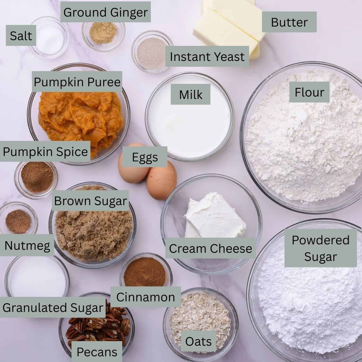 A visual display of the ingredients needed for the Pumpkin Cinnamon Rolls - butter, flour, yeast, milk, eggs, pumpkin puree, nutmeg, ginger, cinnamon, salt, pumpkin spice, granulated sugar, brown sugar, powdered sugar, pecans, oats, and cream cheese - each in their own bowl.