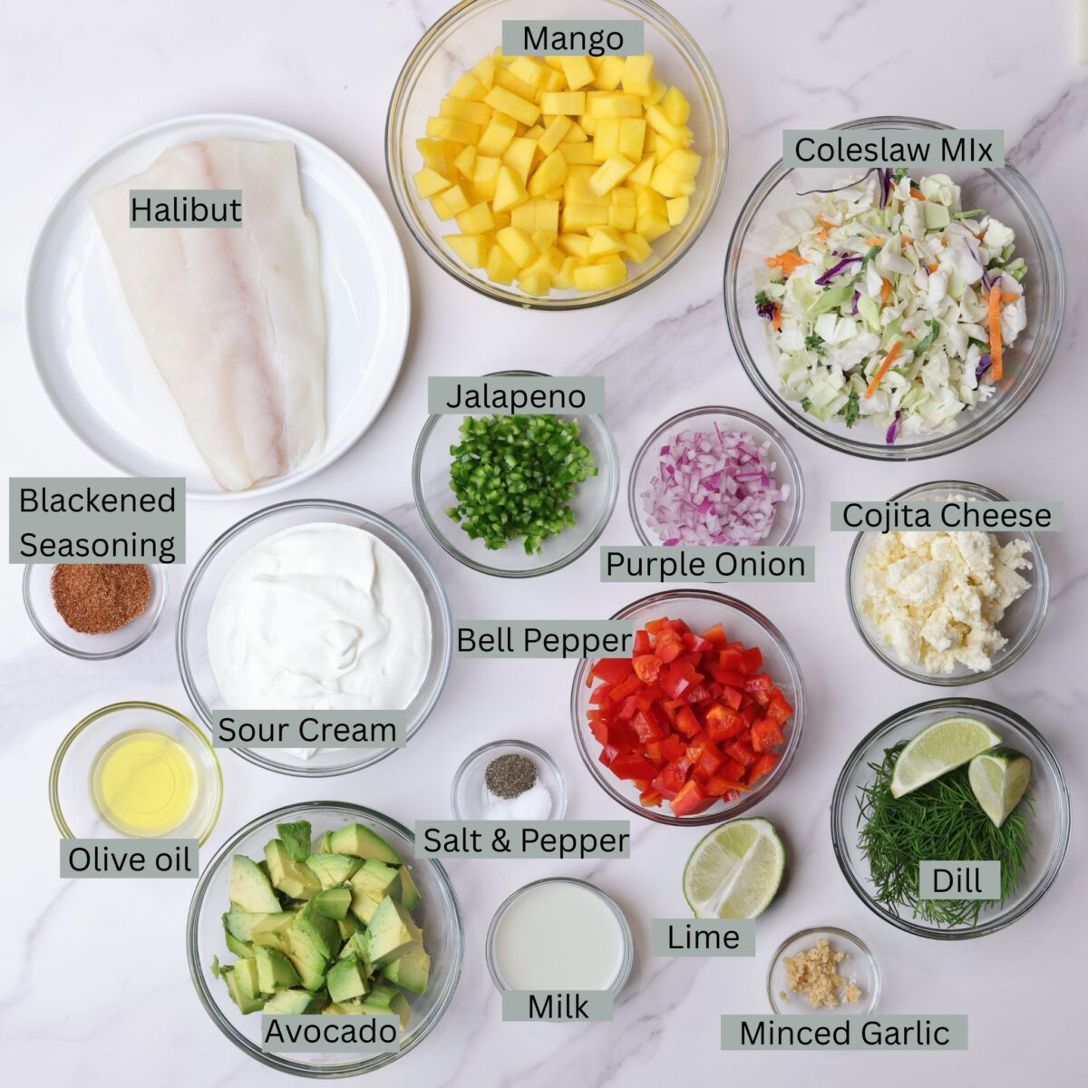 A visual display of the ingredients for blackened halibut tacos - halibut, blackened seasoning, olive oil, sour cream, avocados, red bell peppers, purple onion, jalapeno, mango, shredded cabbage, cojita cheese, fresh dills, minced garlic, milk, limes, salt and pepper - each displayed in their own bowl. 