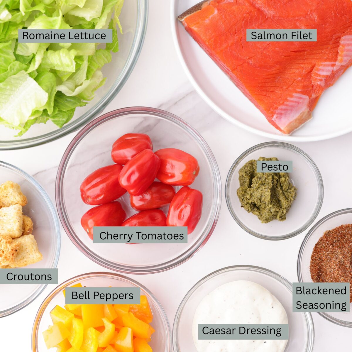 A visual display of the ingredients for Blackened Salmon Caesar Salad - chopped romaine lettuce, 4 ounce raw salmon fillet, croutons, cherry tomatoes, basil pesto, bell peppers, Caesar dressing and blackened seasoning - each in their own individual bowl.