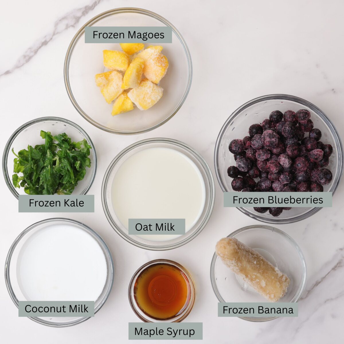 A visual display of the ingredients for the blueberry smoothie without yogurt - frozen mangoes, frozen blueberries, frozen kale, frozen banana, almond milk, coconut milk and maple syrup - each in their own bowl. 