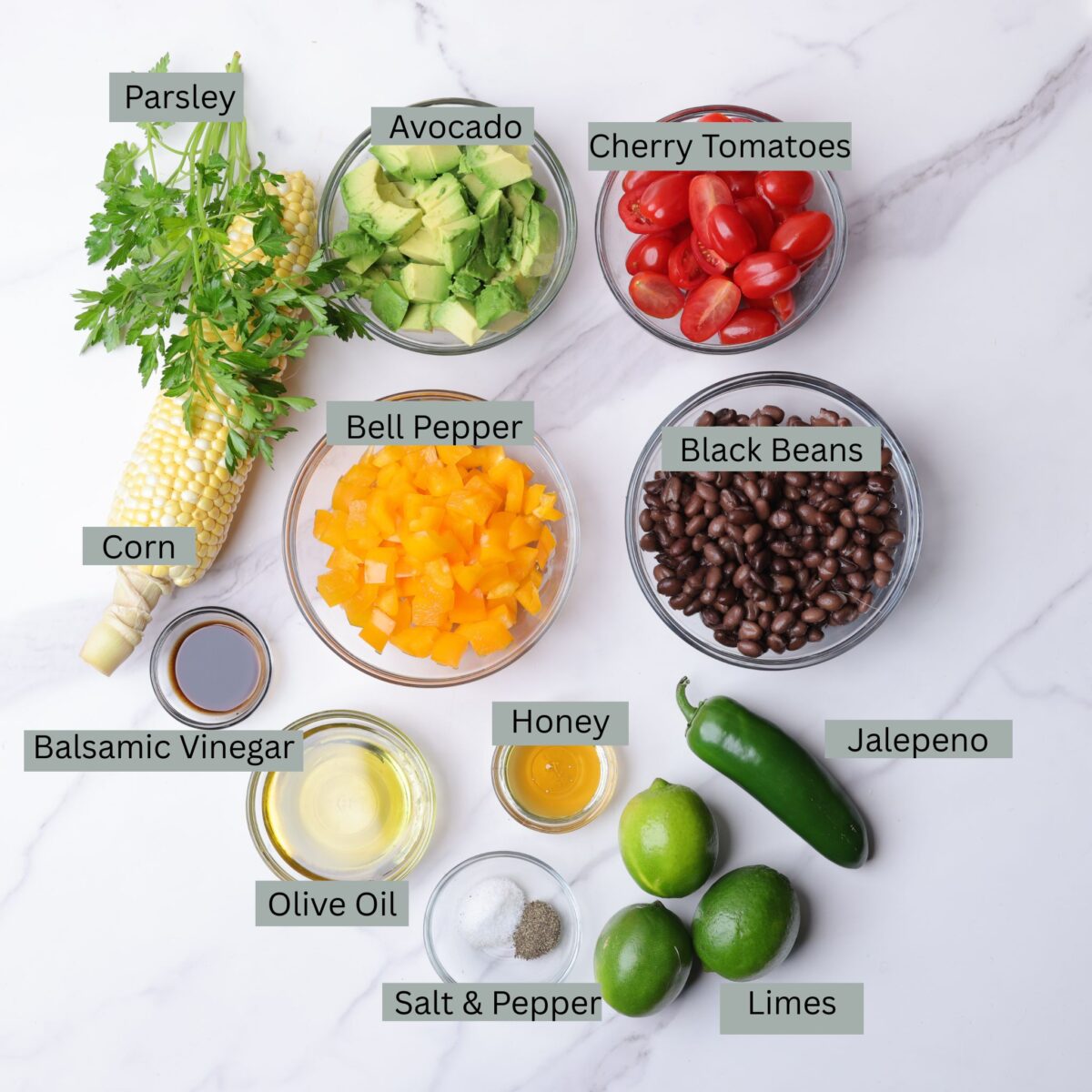 A visual display of the ingredients for the charred corn salad - corn on the cob, parsley, avocado, orange bell pepper, tomatoes, black beans, balsamic vinegar, avocado oil, honey, salt, pepper, limes and a jalapeno.