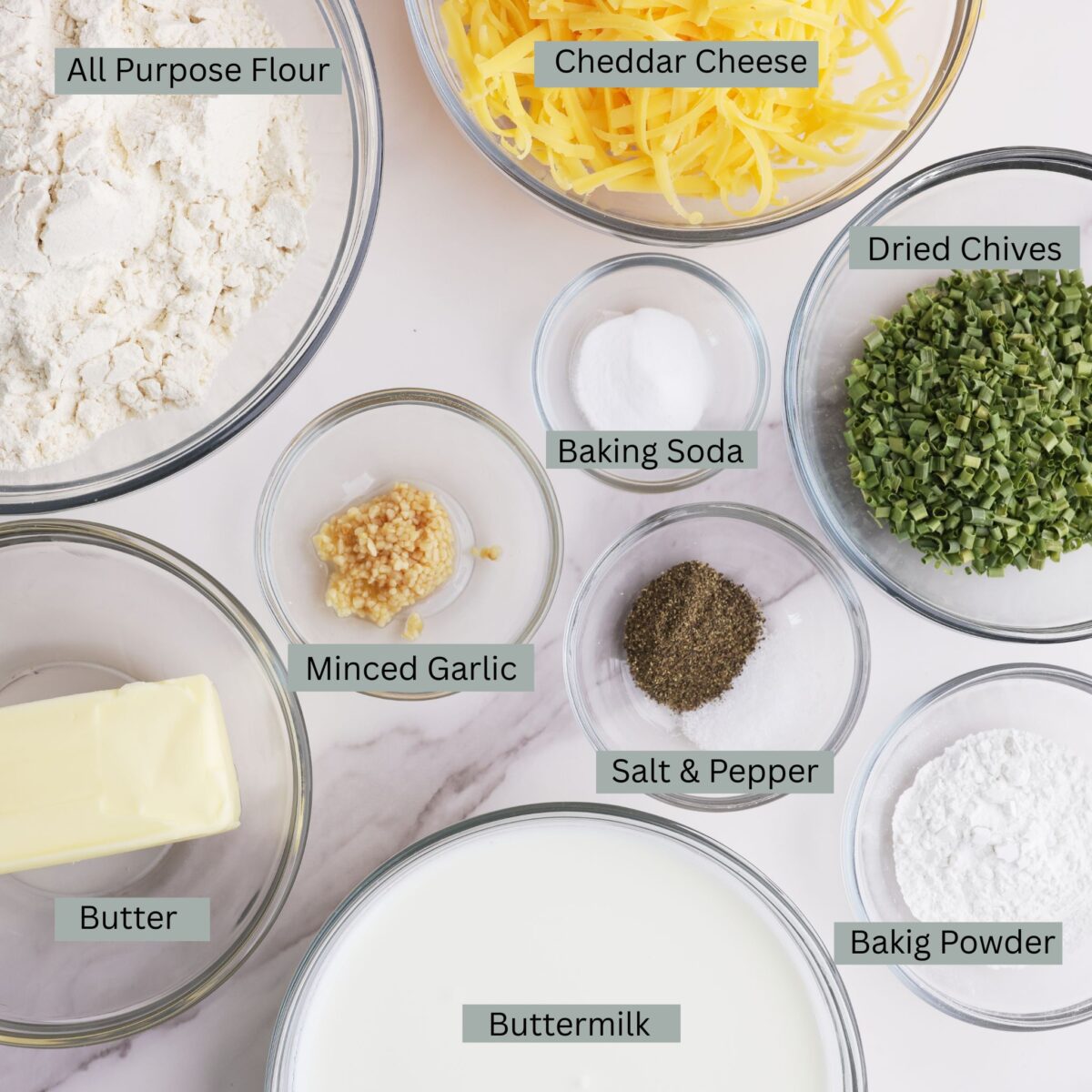 A visual display of the ingredients - flour, dried chives, salt, pepper, garlic, baking powder, baking soda, shredded cheddar cheese, butter and buttermilk - each in their own bowl.