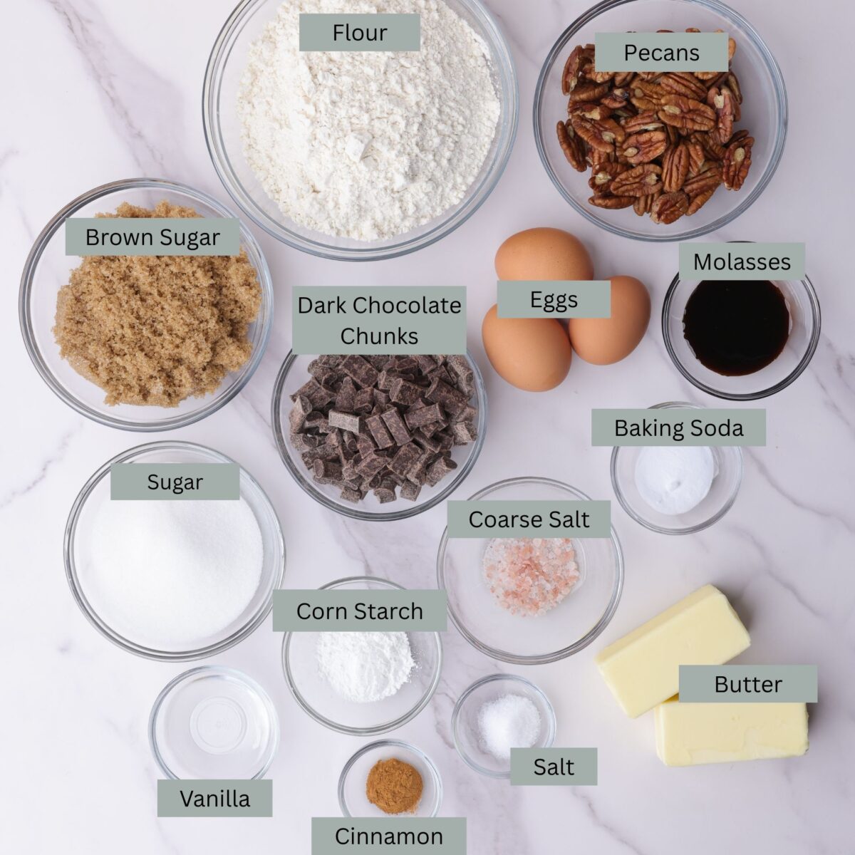 A visual display of the ingredients for Chocolate Chunk Pecan Cookies - flour, pecans, brown sugar, granulated sugar, dark chocolate chunks, eggs, molasses, baking soda, corn starch, coarse salt, salt, butter, cinnamon, and vanilla - each in their own bowl.