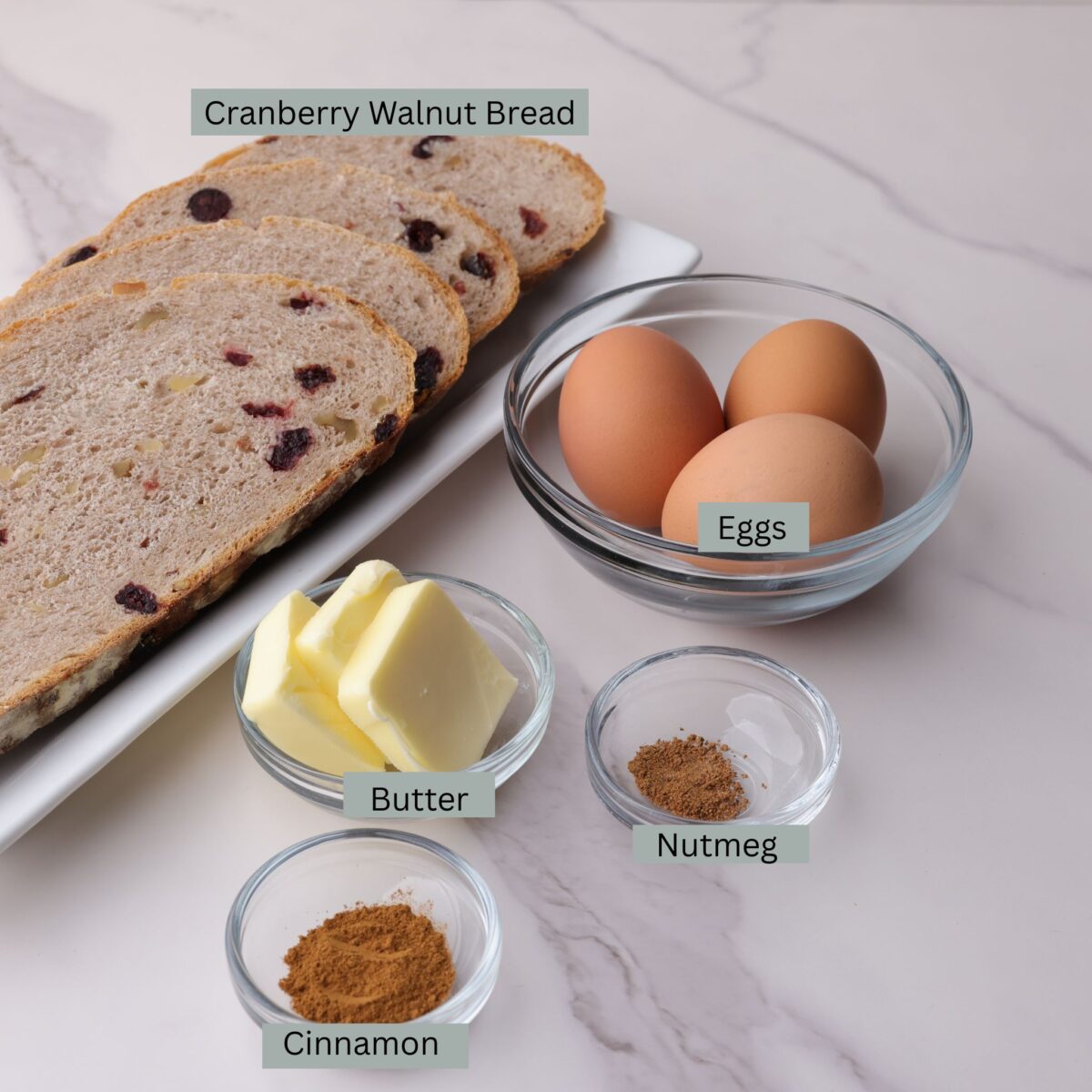 A visual display of the ingredients for cranberry walnut french toast - a plate with four slices of cranberry walnut bread, and eggs, sliced butter, cinnamon and nutmeg each in their own bowl.