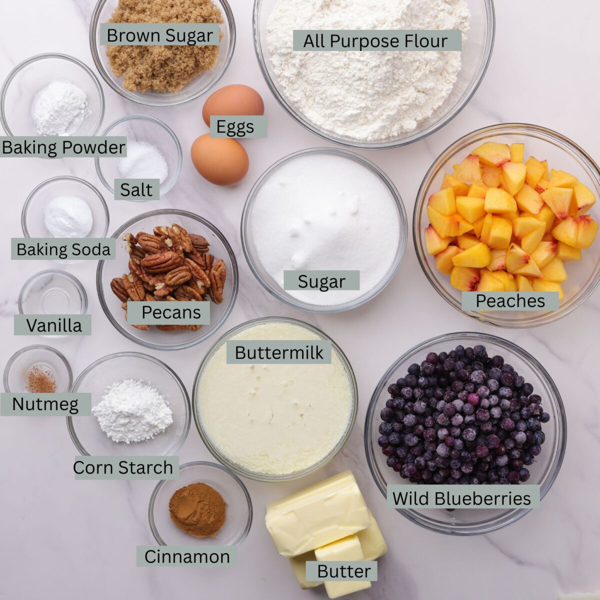 A visual display of the ingredients for the Peach and Blueberry Coffee Cake - flour, brown sugar, granulated sugar, eggs, baking soda, baking powder, salt, cornstarch, vanilla, pecans, buttermilk, cinnamon, nutmeg, butter, peaches and blueberries - each in their own bowl.