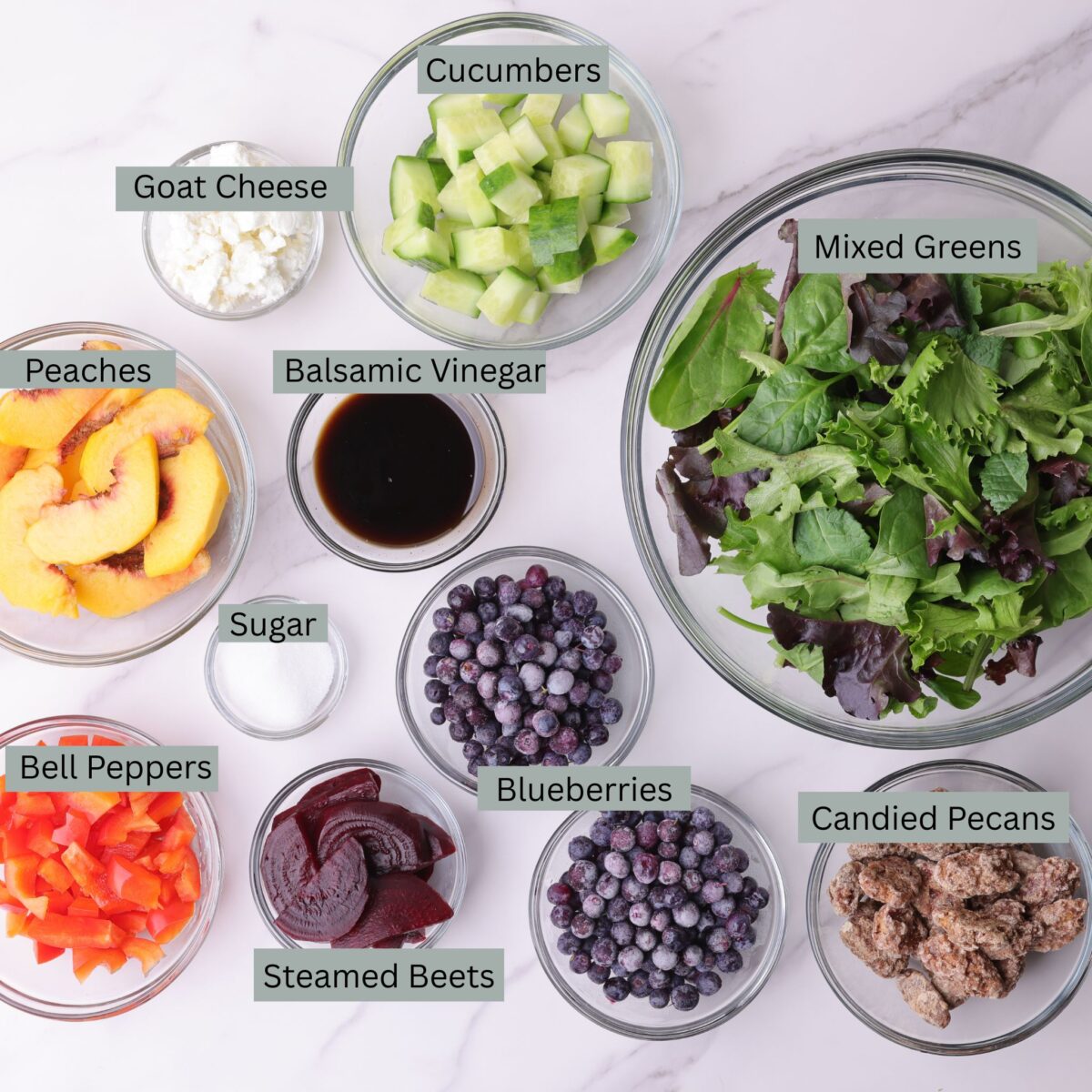 A visual display of the ingredients - mixed greens, cucumbers, goat cheese, peaches, blueberries, red bell peppers, beets, candied pecans, balsamic vinegar and sugar - each in their own bowl. 