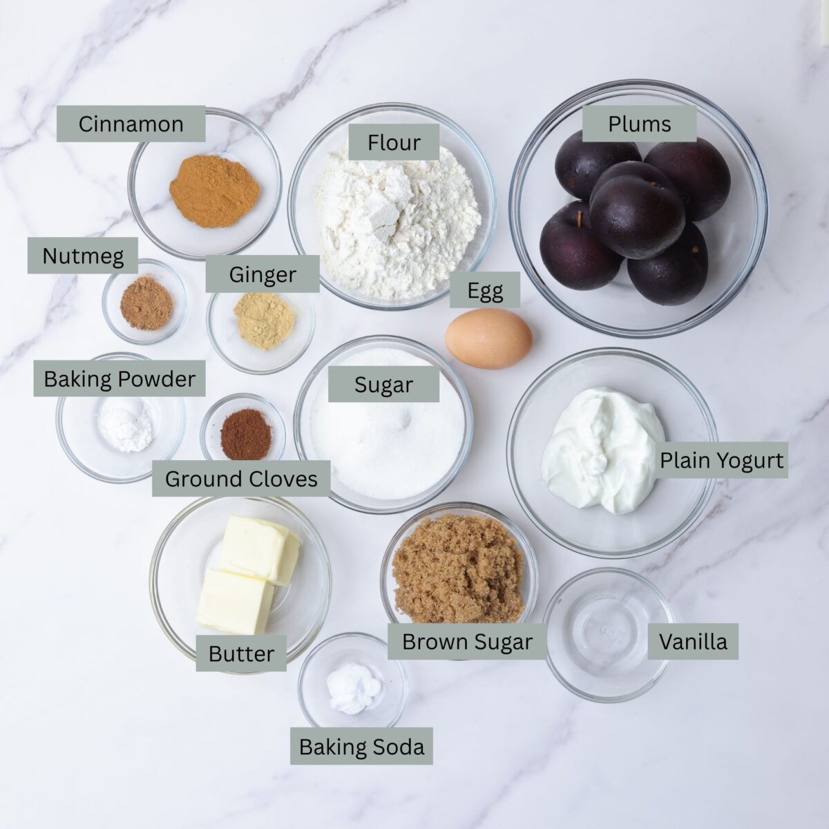 A visual display of the ingredients for the upside down plum cake - plums, flour, granulated sugar, yogurt, butter, brown sugar, vanilla, baking soda, baking powder, an egg, cinnamon, nutmeg, ground ginger, and ground cloves - each in their own bowl.