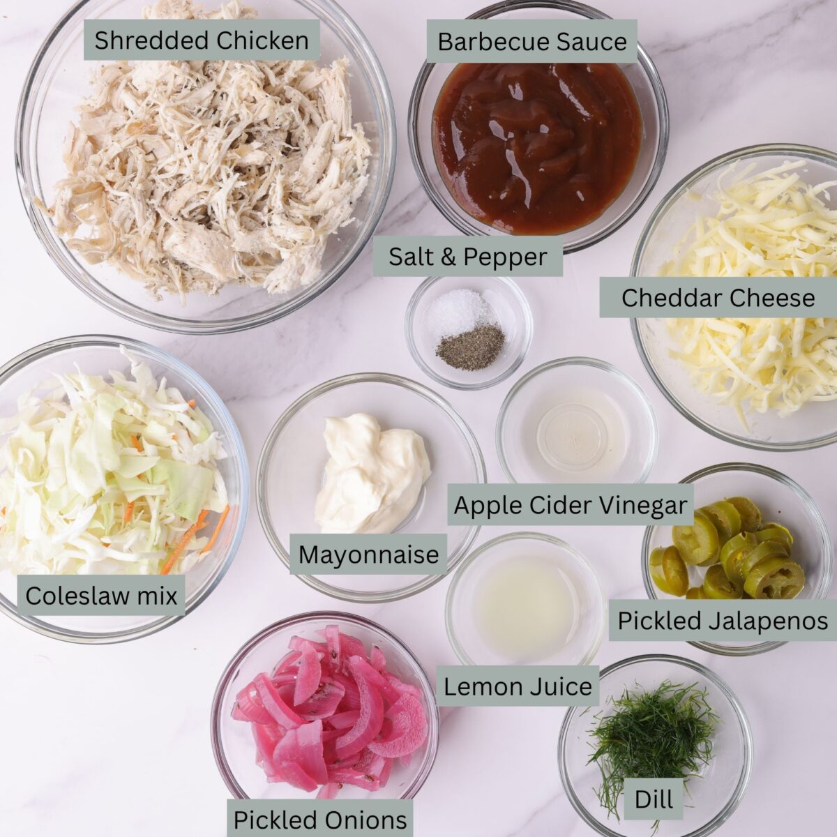 A visual display of the ingredients for the Pulled Chicken Wrap recipe - shredded chicken, barbecue sauce, shredded cheese, salt and pepper, coleslaw, mayonnaise, apple cider vinegar, lemon juice, pickled jalapenos, pickled, purple onions and dill - each in their own bowl.