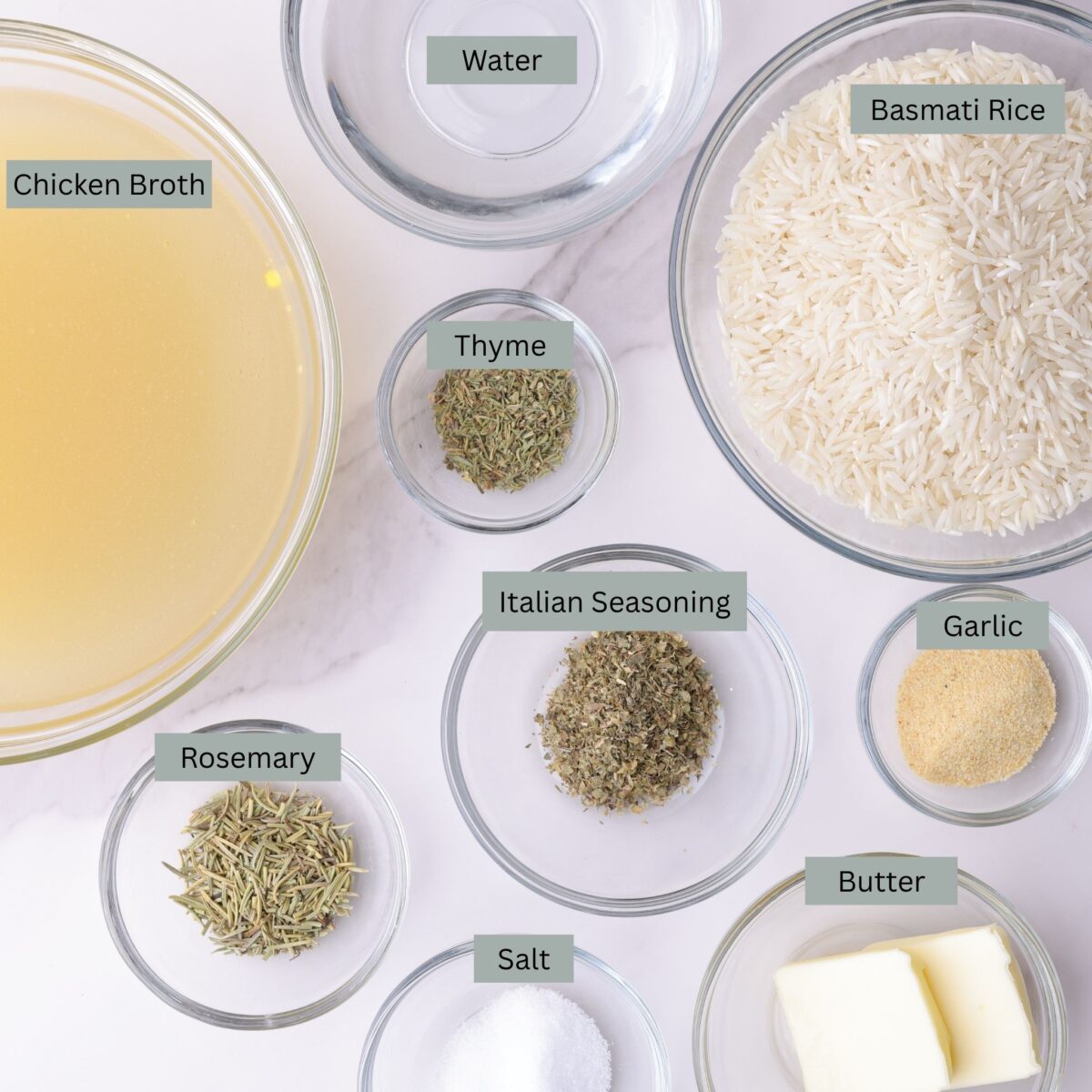 A visual display of the ingredients for the rosemary basmati rice with each ingredient - chicken broth, rice, water, rosemary, thyme, Italian seasoning, garlic powder, salt, and butter - in it's own bowl.