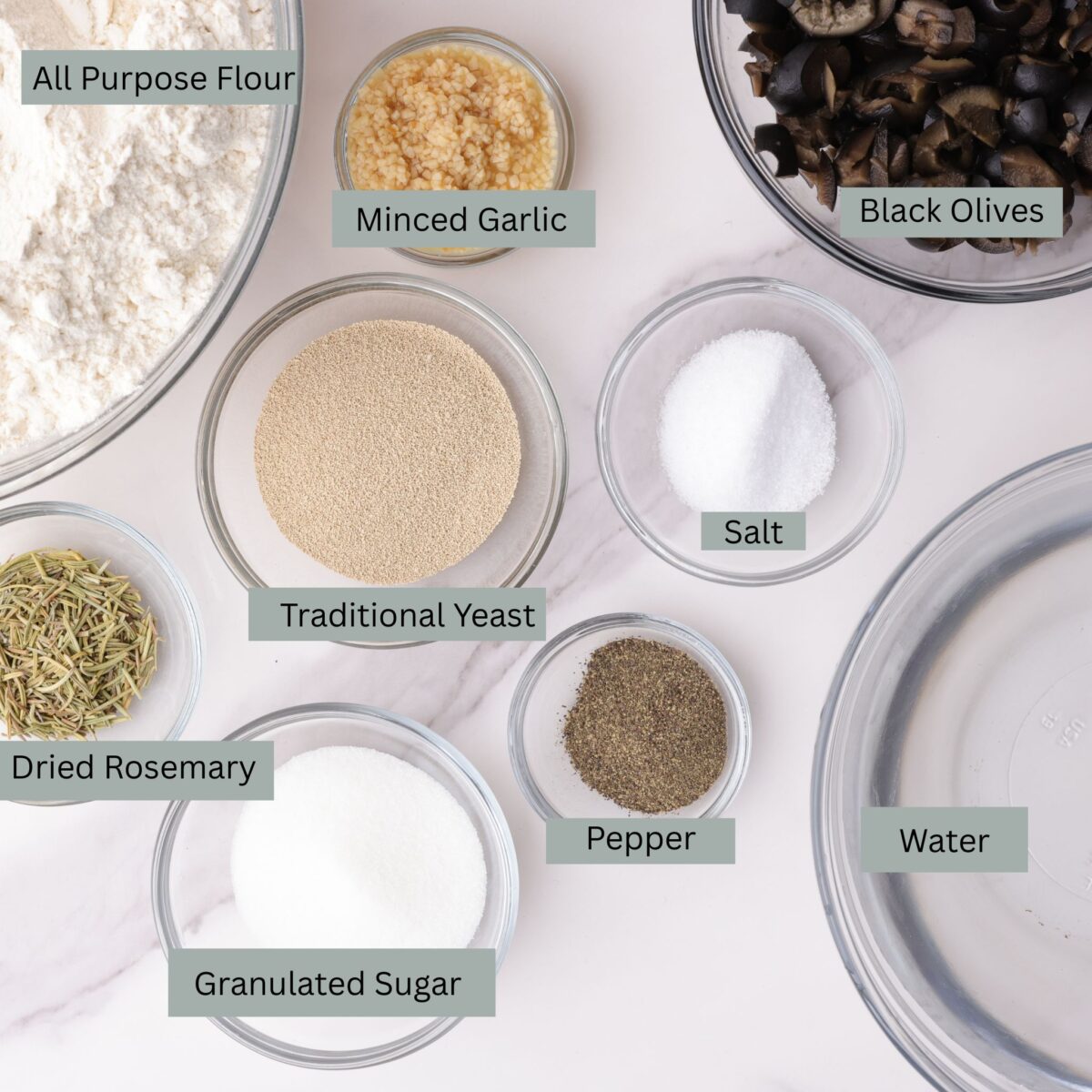 A visual display of the ingredients for the Rosemary Garlic Bread - flour, garlic, olives, yeast, salt, dried rosemary, pepper, sugar, and water - each in their own bowl.