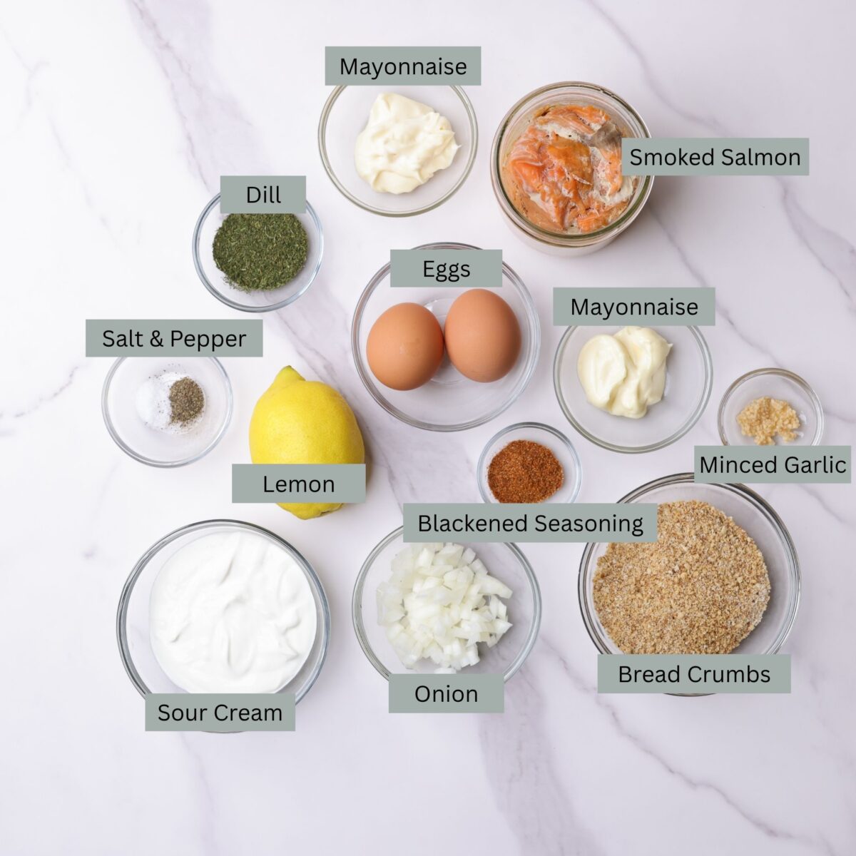 A visual display of the ingredients needed for the salmon meatballs - canned salmon, mayonnaise, eggs, minced garlic, breadcrumbs, chopped onion - as well as the ingredients for the dill sauce - sour cream, salt, pepper, dill and mayonnaise as well as a lemon for juice for both - all displayed in their own bowl.