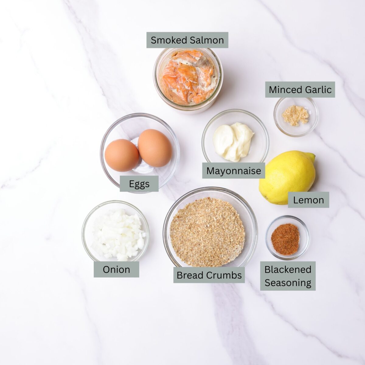 A visual display of the ingredients for Smoked Salmon Patties - smoked salmon, eggs, mayonnaise, minced garlic, chopped onion, breadcrumbs, a fresh lemon and blackened seasoning - each in their own bowl.