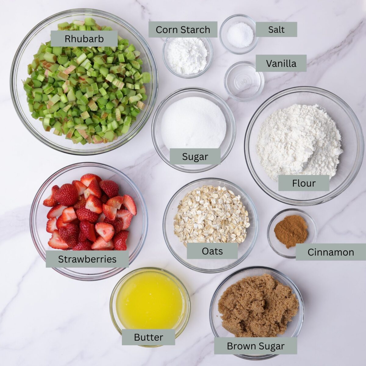 A visual display of the ingredients needed for the crisp - chopped rhubarb, sliced strawberries, corn starch, salt, vanilla, sugar, gluten free flour, gluten free oats, cinnamon, melted butter and brown sugar - each displayed in their own bowl. 