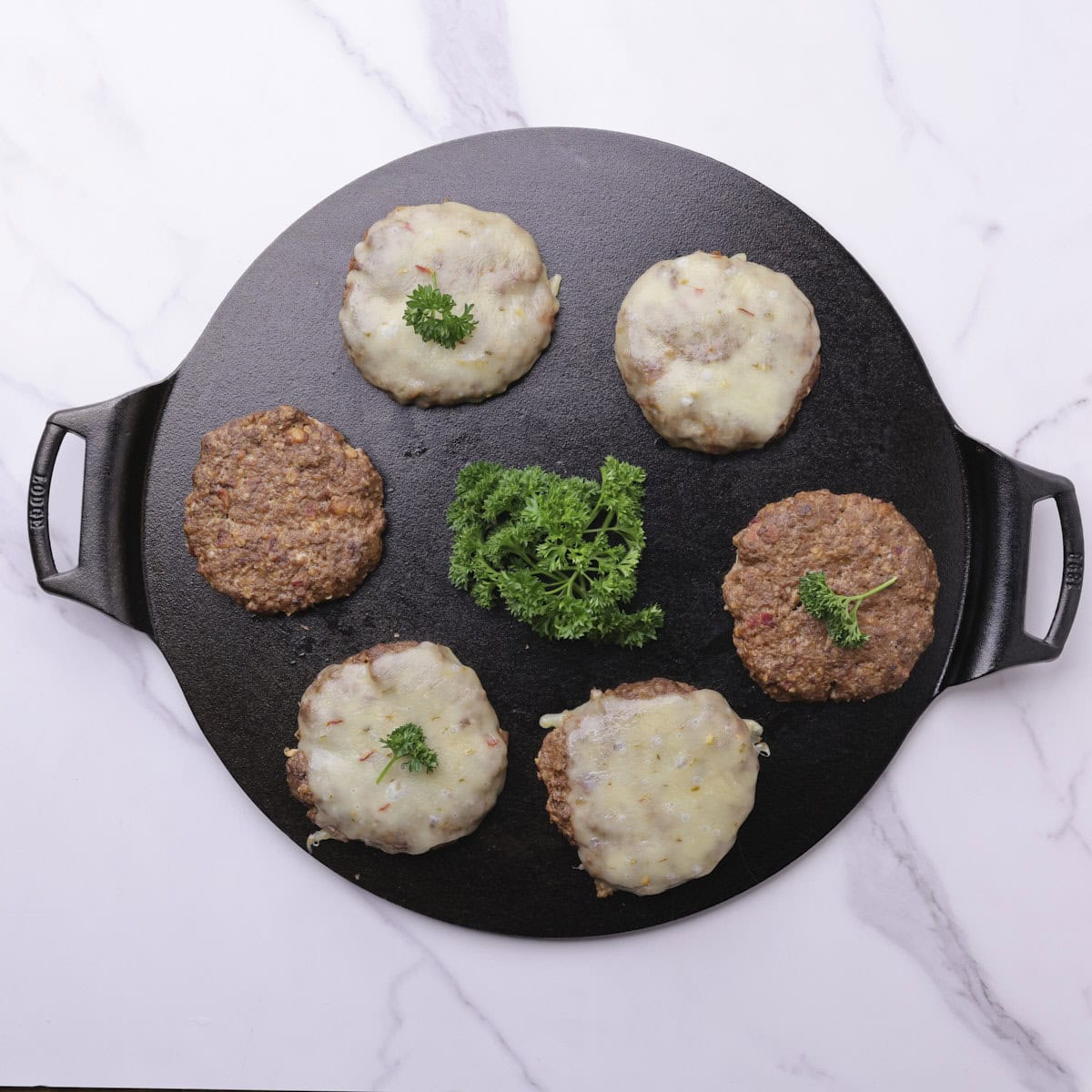 A cast iron pan with 6 cooked moose burger patties on it, 4 with cheese and 2 without and garnished with parsley.
