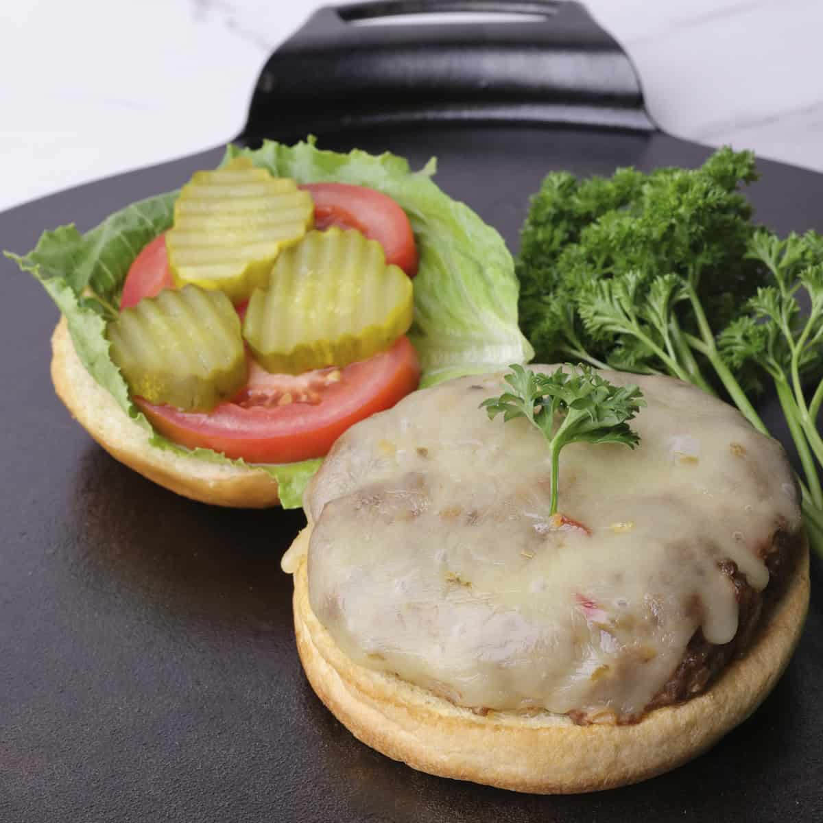 An open faced burger with lettuce, tomato and pickles on one side and the cheese topped burger patty on the other. Displayed with parsley.