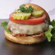A stacked cheese burger with lettuce, bacon, tomato and pickles, garnished with parsley and with the top of the bun leaned against the stack.