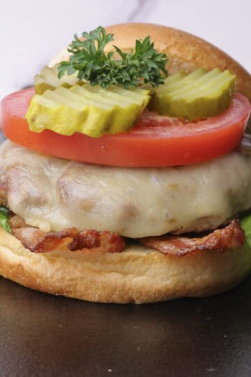 A stacked cheese burger with lettuce, bacon, tomato and pickles, garnished with parsley and with the top of the bun leaned against the stack.