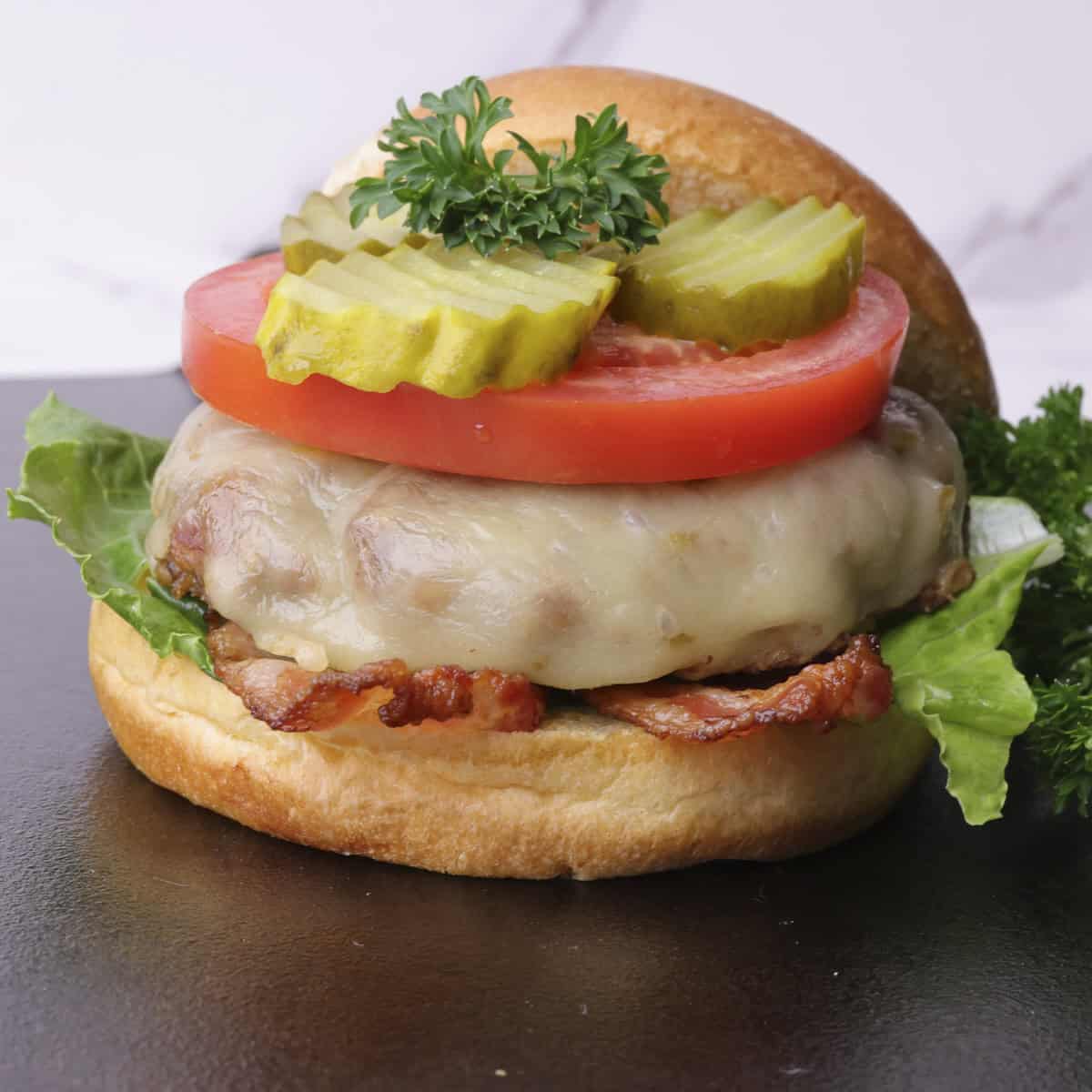 A stacked cheese burger with lettuce, bacon, tomato and pickles, garnished with parsley and with the top of the bun leaned against the stack.