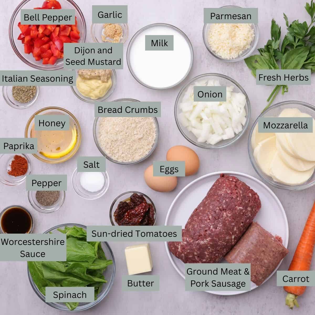 A visual display of the ingredients for the Skillet Meatloaf - bell pepper, garlic, milk, Parmesan, fresh herbs, Italian seasoning, Dijon mustard, seed mustard, paprika, salt, pepper, Worcestershire sauce, honey, breadcrumbs, onions, mozzarella, eggs, sun-dried tomatoes, fresh spinach, butter, ground meat, pork sausage, and a carrot - each in their own bowl.