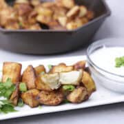 Baked Home Fries displayed on a white rectangular plate with a bowl of dill dip beside and a skillet of home fries in the background.