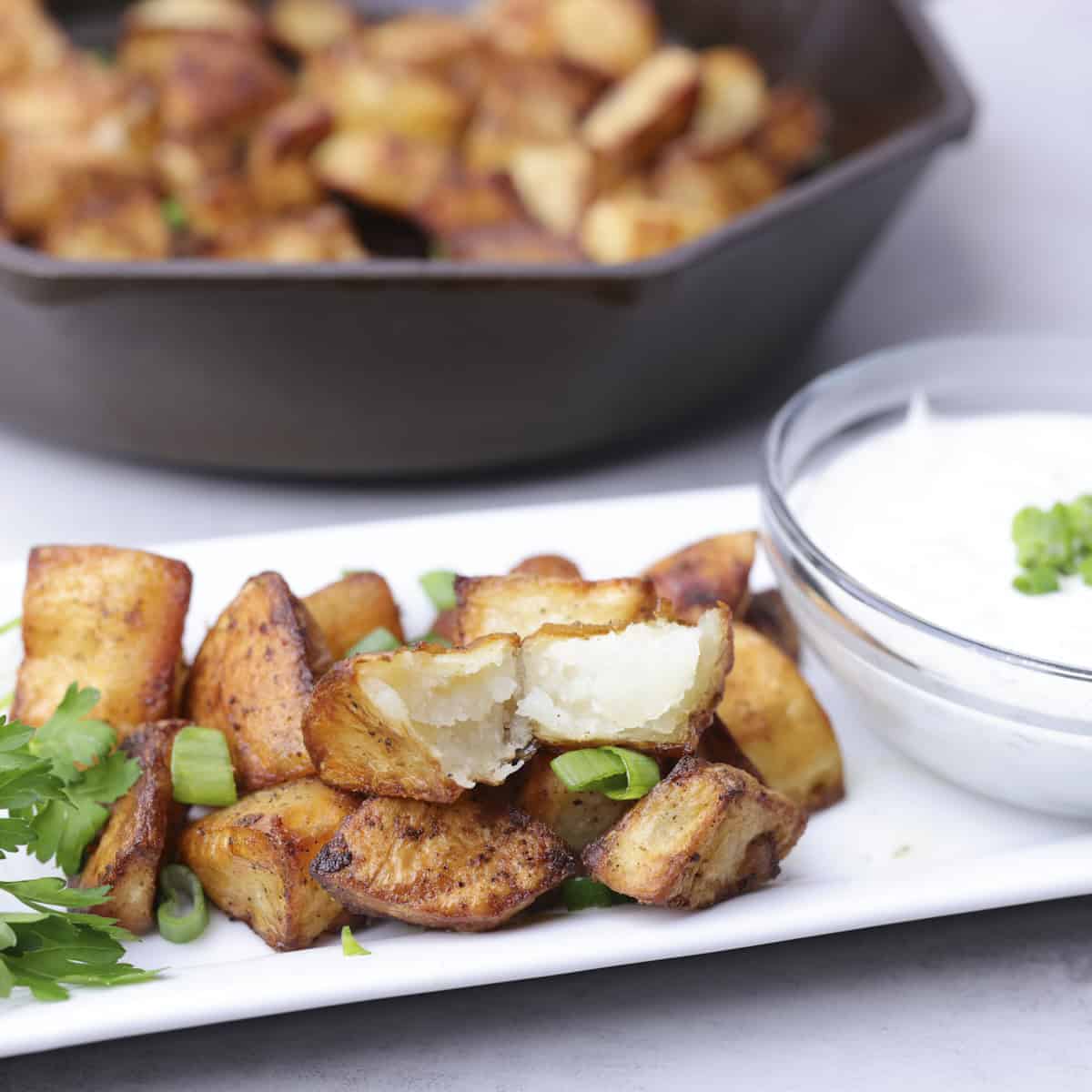 Baked Home Fries displayed on a white rectangular plate with a bowl of dill dip beside and a skillet of home fries in the background.
