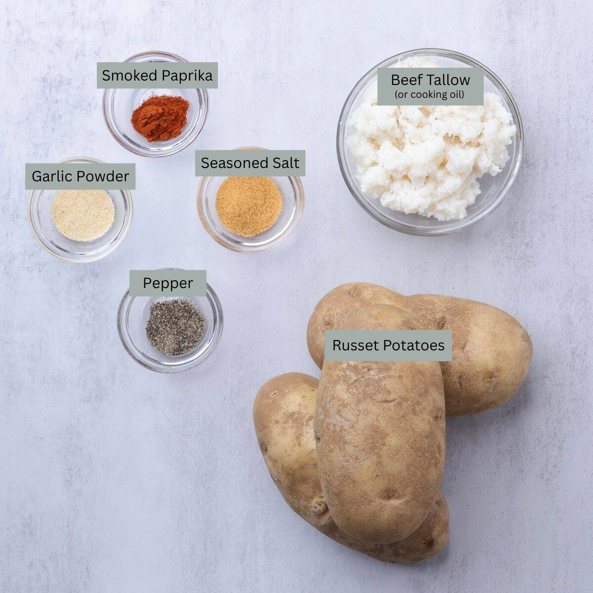 A visual display of the ingredients needed for the Bake Home Fries - beef tallow, russet potatoes, smoked paprika, garlic powder, seasoning salt and pepper - each in their own bowl, except the potatoes.